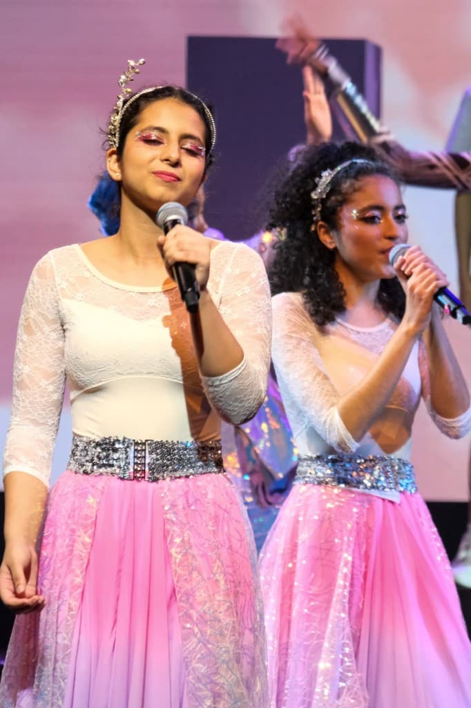 Two young vocalists singing on stage in matching pink skirts, lace tops, and crystal belts