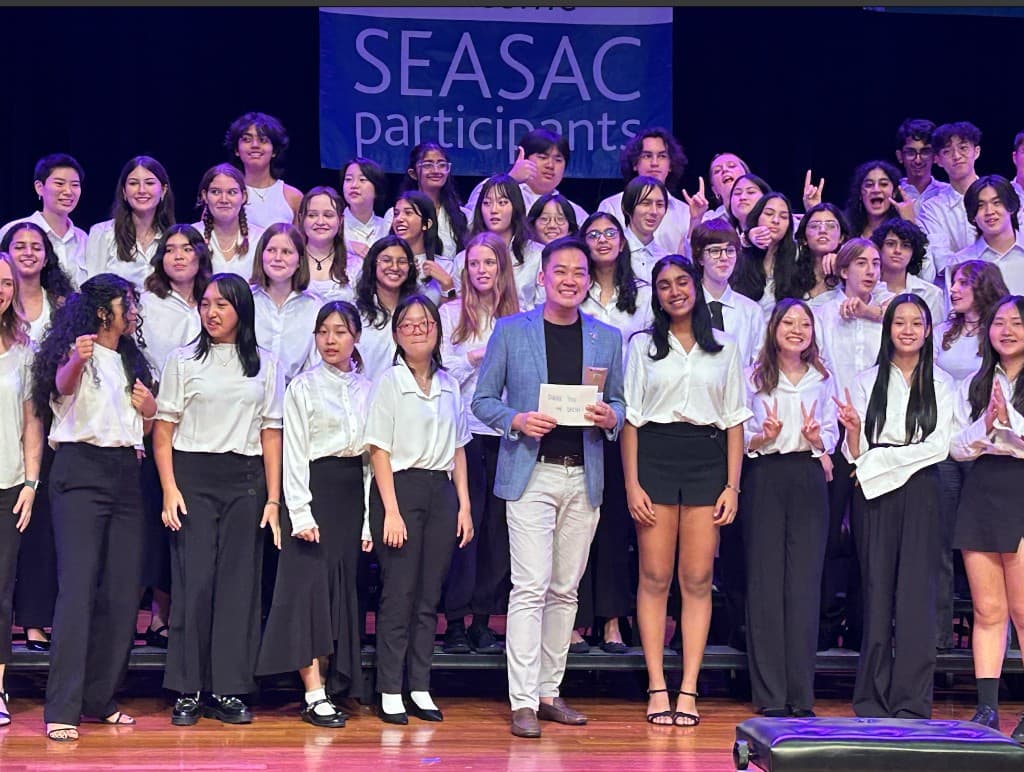 Choir and conductor on stage at SEASAC Music Festival holding a Thank You Mr. Bachelor sign