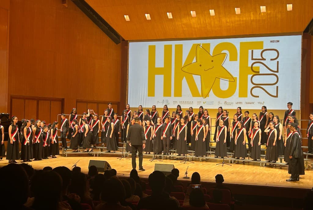 Large youth choir with red-white sashes on risers before HKICF 2025 screen, conductor foreground