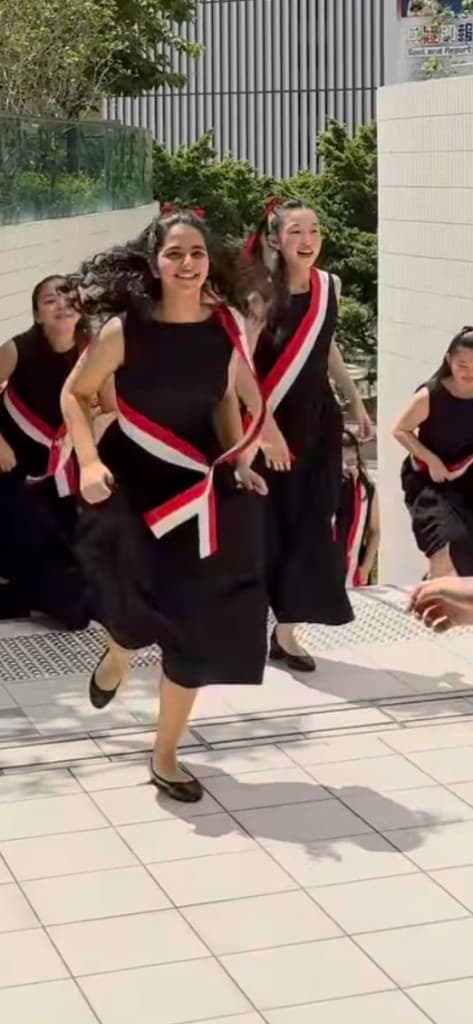 Voices of Singapore choir members in black dresses with red-white sashes running toward the camera on a sunny terrace