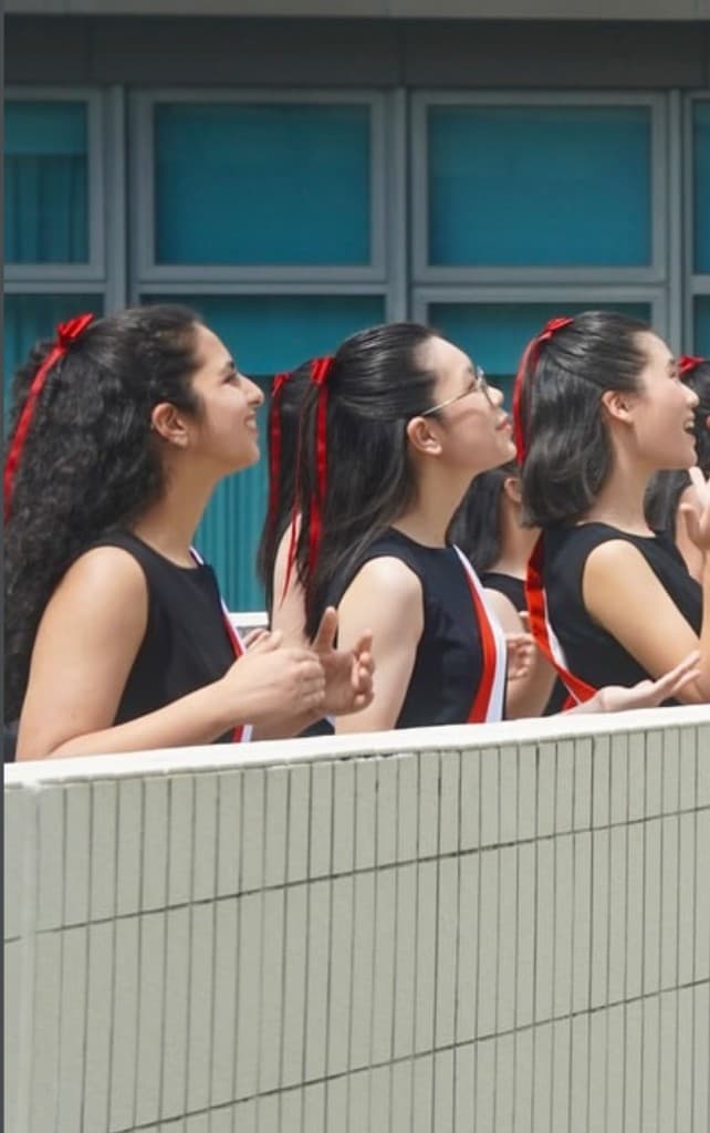 Three choral scholars in matching sashes with red ribbons, smiling upward outdoors