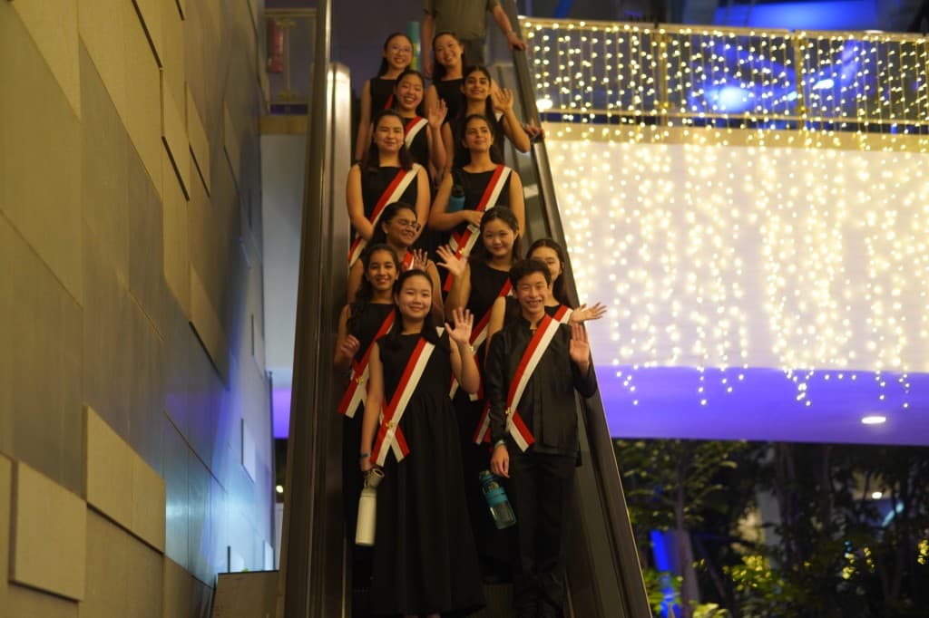 Voices choir in black gowns and red-white sashes smiling and waving on an escalator at night
