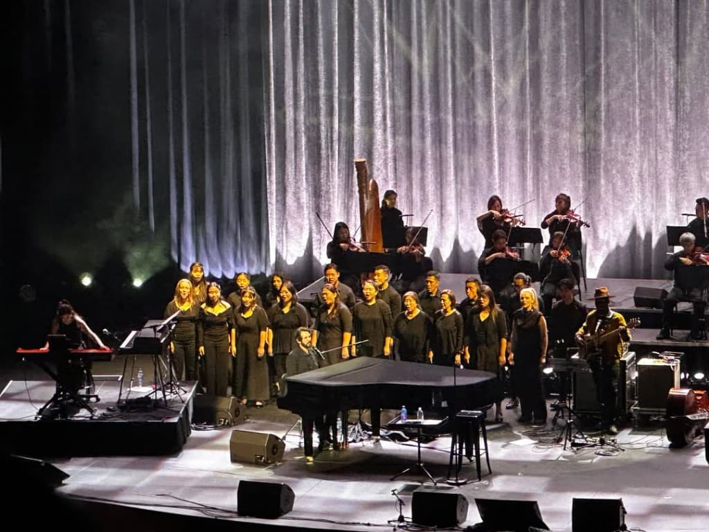 Full orchestral and choral ensemble on concert stage with piano, harp, choir, strings, and guitar